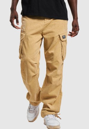 Mann trägt beige Cord-Cargohosen mit Klappentaschen und weißen Sneakern, dargestellt von der Taille bis zu den Füßen vor einem einfarbigen Hintergrund.