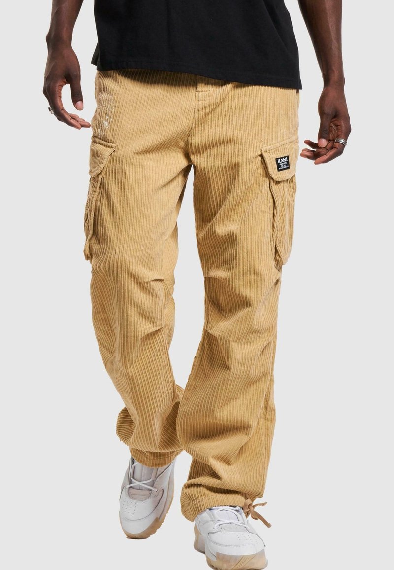 Homme portant un pantalon cargo en velours côtelé beige avec des poches à rabat et des baskets blanches, montré de la taille aux pieds sur un fond uni.