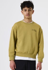 Sudadera de color mostaza con cuello redondo y puños de canalé. Presenta un pequeño estampado negro en el pecho y se lleva sobre una camisa blanca.