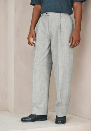 Mann trägt graue Plissee-Hose, schwarze Loafers und ein dunkelblaues Kurzarmhemd und steht an einer beigen Wand.