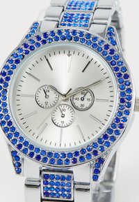 Reloj plateado con un bisel azul adornado con piedras de strass. Presenta tres sub-diales y una pulsera metálica lisa con un acabado pulido.