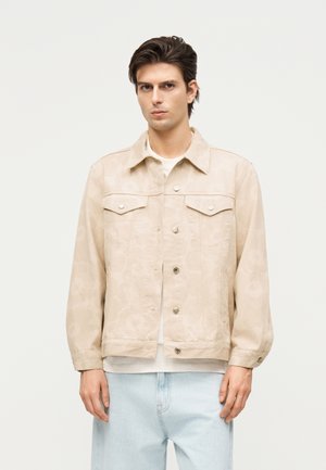 Fiatal férfi bézs színű, gombos zakóban, fehér ing és világoskék farmer felett, egyszínű fehér háttér előtt áll.