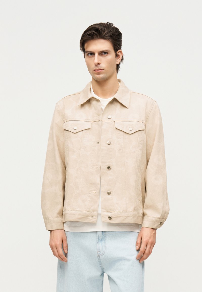 Νέος άνδρας που φορεί μια μπεζ σακάκι με κουμπιά πάνω από ένα λευκό πουκάμισο και ανοιχτό μπλε τζιν, που στέκεται μπροστά σε απλό λευκό φόντο.