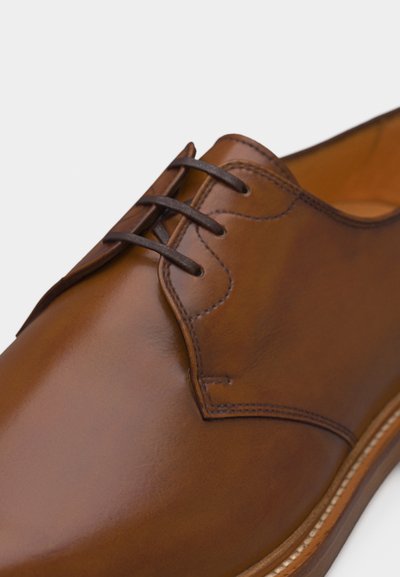 Καφέ δερμάτινο παπούτσι με σκούρα καφέ κορδόνια, λεπτομερείς ραφές και λεία γυαλιστερή επιφάνεια, φωτογραφημένο σε ανοιχτό φόντο.