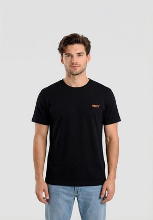 Joven con cabello castaño que lleva una camiseta negra con la palabra "HÉROE" y jeans azul claro, de pie frente a un fondo gris liso.