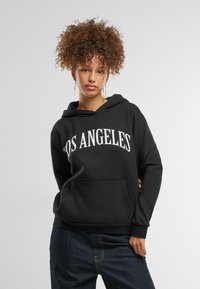 Fekete kapucnis pulóver, az elején vastag fehér betűkkel "LOS ANGELES" felirattal, elülső zsebével és laza szabással, sötét farmernadrággal párosítva.