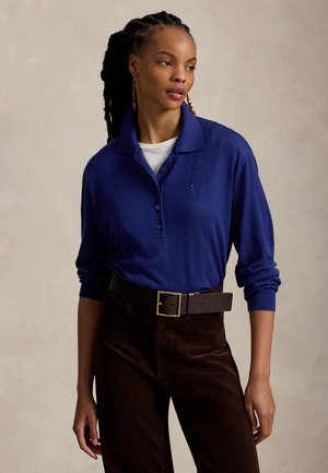 Femme aux cheveux tressés portant un polo à manches longues bleu, un sous-vêtement blanc, un pantalon marron et une large ceinture marron avec une boucle dorée.