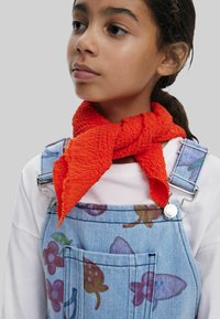 Sciarpa arancione con texture annodata intorno al collo, indossata con una salopette in denim blu con motivi floreali multicolori e dettagli in metallo argentato.