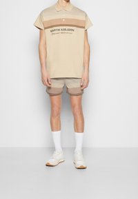 Beige pikétröja med horisontella ljusbruna och mörkbruna ränder, i kombination med matchande shorts. Vita sneakers och vita strumpor i ankellängd.