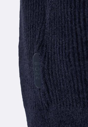 Mörkblå stickad tyg med ett texturerat mönster. Inkluderar en liten svart etikett med präglad text. Fokus på material och designdetaljer.