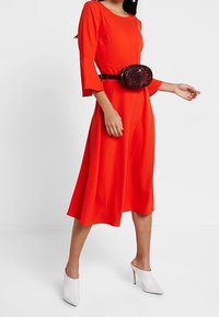 Robe rouge mi-longue aux manches trois-quarts, en tissu lisse ; associée à un sac ceinture en cuir serpent noir et rouge et des mules blanches à talons.