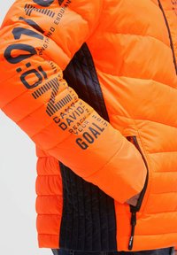 Orange puffer jacket with black side panels, featuring prominent text and patterns. Zippered pocket on the right side adds functionality.