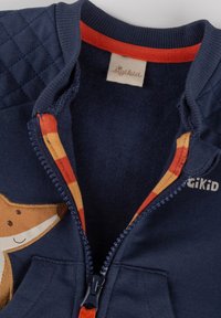 Marineblauer Zip-Jacke mit gesteppten Schultern, orange- und gelb kariertem Innenfutter. Mit einer Applikation eines braunen Fuchses und dem "sigikid"-Logo auf der Brust.