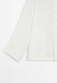 Chemise blanche en lin à manches longues avec fermeture à boutons sur le devant et une poche latérale visible sur un fond uni.