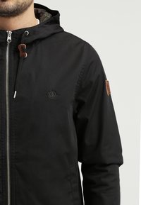 Czarna kurtka z kapturem z przednim zamkiem błyskawicznym, z logo na piersi, beżowymi skórzanymi akcentami i wzorzystą podszewką wewnętrzną.