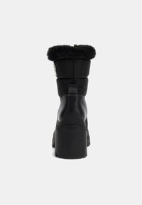 Botte d'hiver noire mi-mollet avec garniture en fourrure, talon épais, semelle robuste et logo Guess sur le talon, vue de l'arrière.