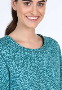 Tropfengrünes Oberteil mit einem runden Ausschnitt und einem strukturierten Design aus ineinandergreifenden Blattmotiven. Der Stoff wirkt weich und glatt.