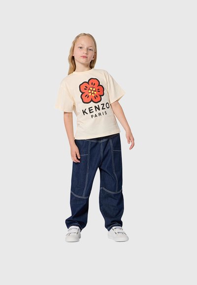 Meisje dat staat, draagt een beige T-shirt met een grote rode bloem en de tekst "Kenzo Paris", donkerblauwe spijkerbroek en witte sneakers.