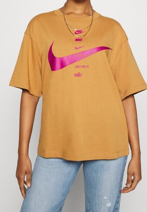 Osoba nosząca beżowy t-shirt Nike z dużym różowym logo typu swoosh i napisem, w połączeniu z jasnoniebieskimi dżinsami i warstwowymi złotymi łańcuszkami.