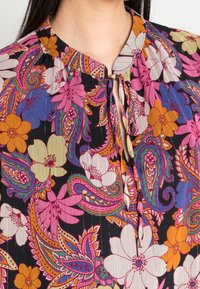 Blouse multicolore avec un fond noir orné de motifs floraux et cachemire en rose, violet, orange et crème. Se noue au niveau du cou.