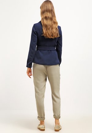 Femme aux longs cheveux ondulés portant une veste ceinturée marine, un pantalon beige retroussé et des mocassins dorés, debout devant un fond blanc uni.