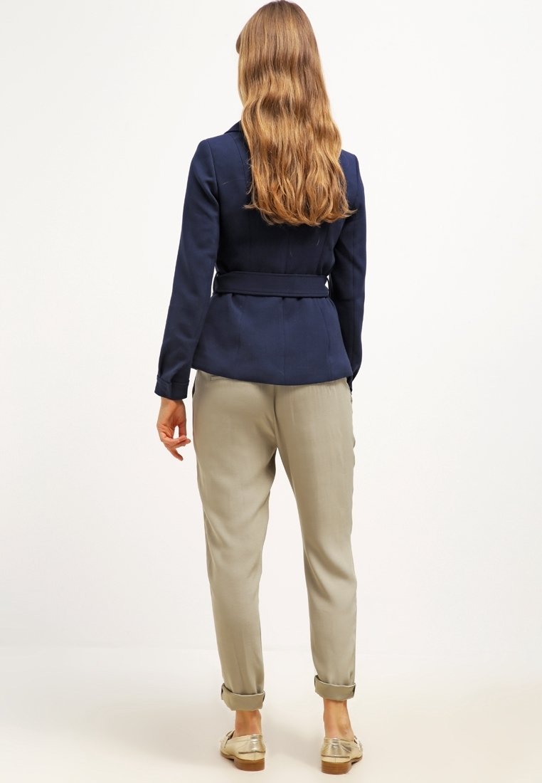 Femme aux longs cheveux ondulés portant une veste ceinturée marine, un pantalon beige retroussé et des mocassins dorés, debout devant un fond blanc uni.