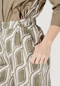 Pantaloni a vita alta con un motivo astratto verde e bianco, caratterizzati da pieghe, una cintura con una chiusura in metallo e un tessuto morbido e testurizzato.