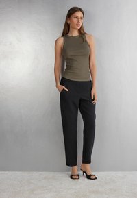 Top de tirantes acanalado en oliva combinado con pantalones negros ajustados. La modelo lleva sandalias de tacón negras. Un fondo gris suave realza el atuendo.