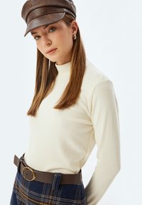Beige gerippter Rollkragenpullover kombiniert mit hoch taillierten navyblauen Karohosen. Braune Lederkappe und Gürtel mit einer runden goldenen Schnalle.