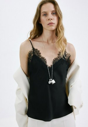 Femme aux cheveux blonds ondulés portant un caraco noir bordé de dentelle, une veste blanche drapée sur les bras et un long collier argenté avec des breloques géométriques.