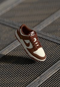 Smeđe i bež Nike tenisice s vezicama na teksturiranoj metalnoj površini, prikazane bočno i odozgo s vidljivim detaljima logotipa.