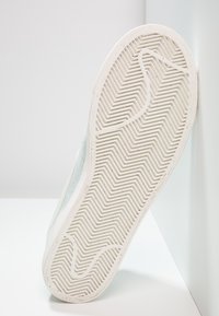 La semelle de chaussure en caoutchouc blanc présente un motif de crampons en chevrons. Le côté montre un daim bleu clair doux avec un design épuré et des accents minimaux.