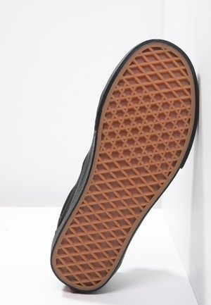 Semelle de chaussure avec motif gaufré en caoutchouc marron et bord noir, positionnée devant un fond blanc d’angle.