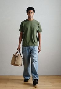 Camiseta verde-oliva, jeans soltos de ganga azul e sapatos pretos. Carrega uma mochila de lona cor de castanho claro com alças escuras e detalhes castanhos.