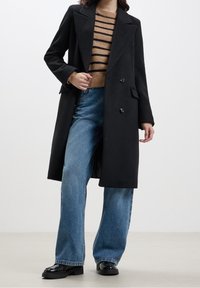 Cappotto nero doppiopetto con revers, indossato sopra un maglione a righe marroni e jeans larghi azzurri. Scarpe nere lucide.