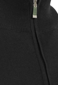 Primo piano di un tessuto di maglia nera con una cerniera metallica e un tirante della cerniera verde stampato con un logo.