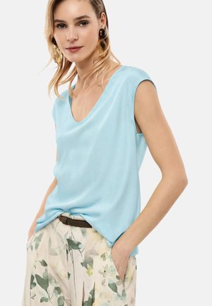 Donna con top azzurro senza maniche, pantaloni a gamba larga a fiori con cintura, mani nelle tasche e orecchini vistosi marroni e dorati.