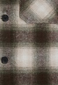 Braun- und beige-kariertes Flanellgewebe mit Fischgrätenstruktur, ausgestattet mit dunklen Knöpfen und einer aufgesetzten Tasche in der oberen linken Ecke.
