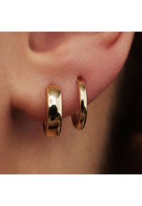 Goldene Creolen mit glanzendem Finish, die eine glatte, runde Form und einen nahtlosen Verschluss aufweisen, die am Ohr eines Modells präsentiert werden.
