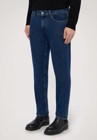 Tmavě modré džíny z denimu se s rovnym střihem, nabízející přední kapsy, zip a knoflíkové zapínání. Nosené s černými koženými kotníkovými botami.