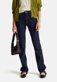 Jeans de mezclilla azul oscuro con costuras visibles, combinados con una blusa clara y un cárdigan verde. Se incluye un bolso negro pequeño y zapatos negros.