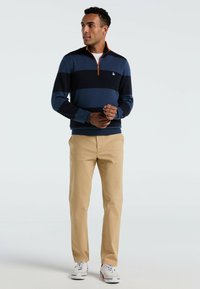 Marineblauw en lichtblauw gestreepte trui met een halve rits en oranje accenten, gecombineerd met beige broeken en witte sneakers.