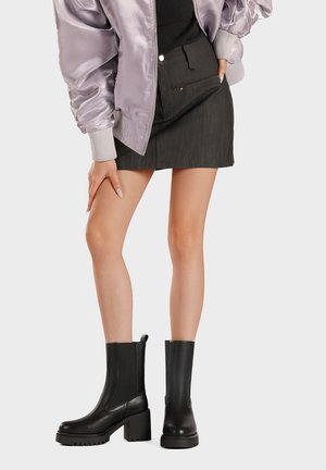 Donna con giacca bomber in raso viola, minigonna nera e stivali neri con tacco a blocco a metà polpaccio, in piedi davanti a uno sfondo semplice.