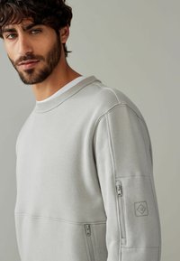 Lys grå sweatshirt med ribbet krave, sidelommer og tekstureret stof. Har logo-patch på ærmet for detalje.