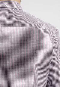 Rückansicht einer Person, die ein weißes und burgunderfarbenes, klein karriertes Langarmhemd mit einem Knopf am Kragen trägt.