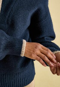 Navyfarbener Wollpullover mit strukturiertem Strick, der cremefarbene Rippenbündchen aufweist. Nahaufnahme von Händen, die die Ärmel vor einem hellen Hintergrund anpassen.