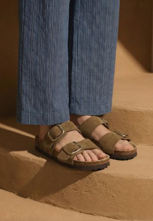 Picioare care poartă sandale maro din piele întoarsă cu catarame mari și pantaloni albaștri cu dungi fine, stând pe trepte bej texturate.