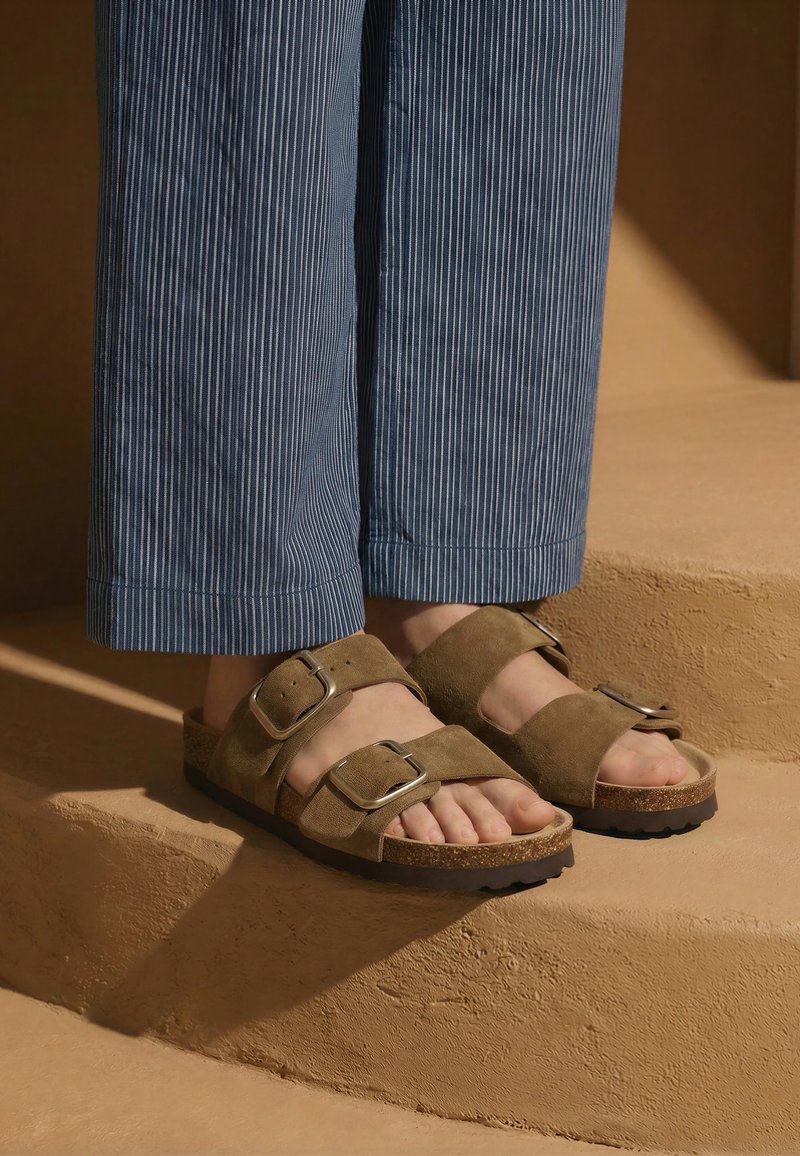 Pieds portant des sandales en daim marron avec de grandes boucles et un pantalon bleu à fines rayures, debout sur des marches beige texturées.