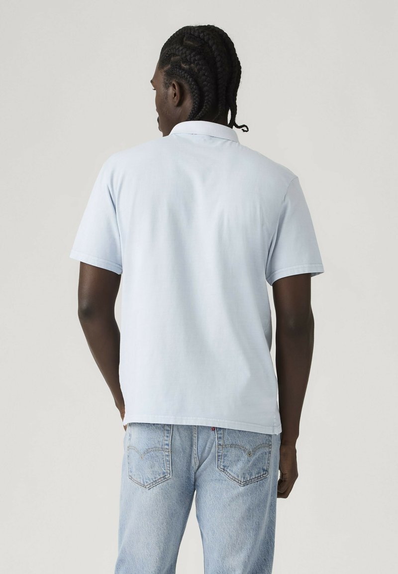 Hombre con cabello trenzado, vistiendo una camiseta polo de manga corta de color azul claro y jeans azul claro, de pie de espaldas contra un fondo liso.