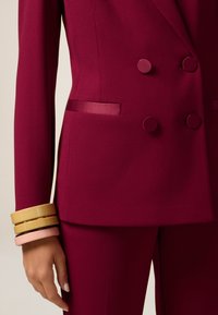 Giacca su misura fucsia con bottoni tondeggianti, tasche laterali discrete e rifiniture in satin. Accentuata da bracciali colorati impilati al polso.